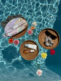 Trousse à maillot de bain Owaïna avec ses accessoires de plage, crème solaire, chouchou et lunettes de soleil. Présenté flottant au dessus d’une piscine sur des plateau en osier et des fleurs.