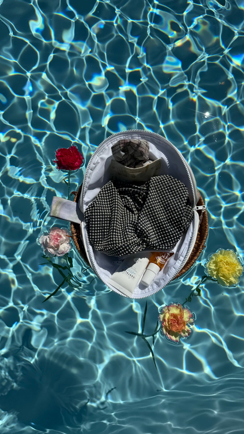 Trousse à maillot de bain ouverte avec ses accessoires de plage. Lunette de soleil, chouchou, crème solaire. Présenté flottant au dessus d’une piscine sur un panier en osier et des fleurs.